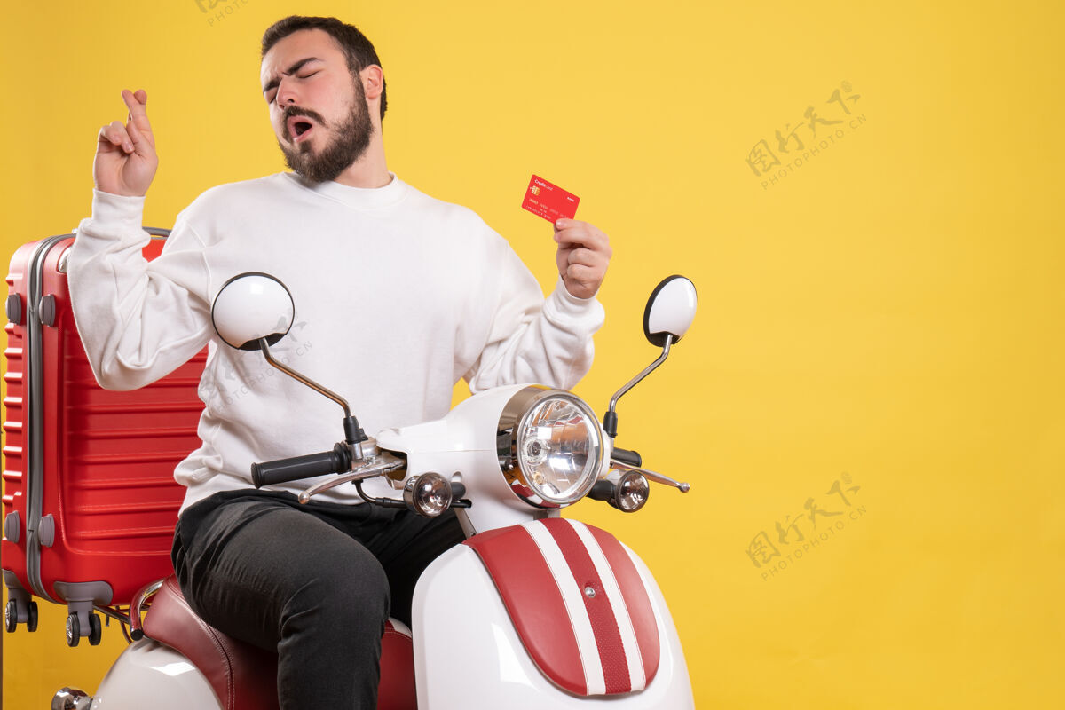 摩托车年轻的梦幻旅行男子坐在摩托车上 手提箱上拿着银行卡 手指交叉在孤立的黄色背景上摩托车男子旅游黄色