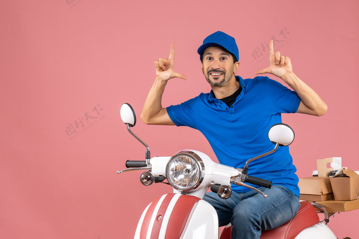 背景前视图微笑和快乐的信使家伙戴着帽子坐在粉彩桃色背景上踏板车微笑踏板车人物