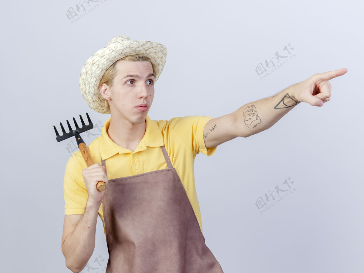 年轻人年轻的园丁 穿着连体衣 戴着帽子 拿着小耙子 用食指指着旁边的东西 表情严肃脸目录男人