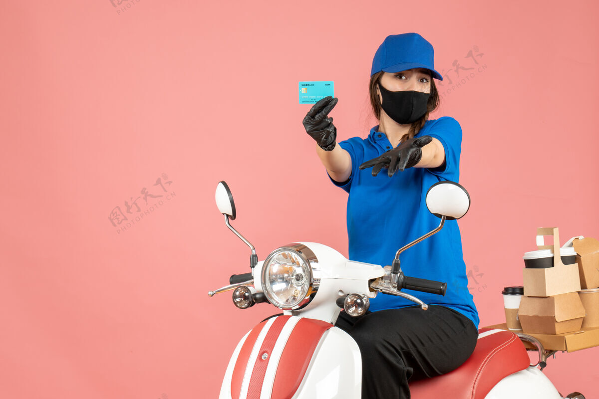 摩托车带着医用面罩和手套的快递员女孩坐在滑板车上 拿着银行卡 在柔和的桃色背景下递送订单背景医疗穿
