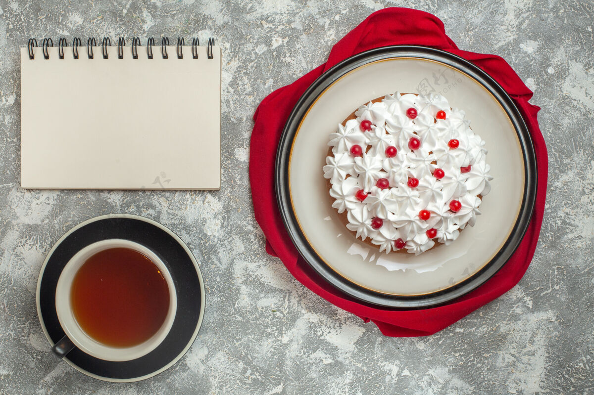 背景上图：美味的奶油蛋糕装饰着红色毛巾上的水果和一杯红茶 旁边放着冰背景笔记本笔记本过滤器杯子