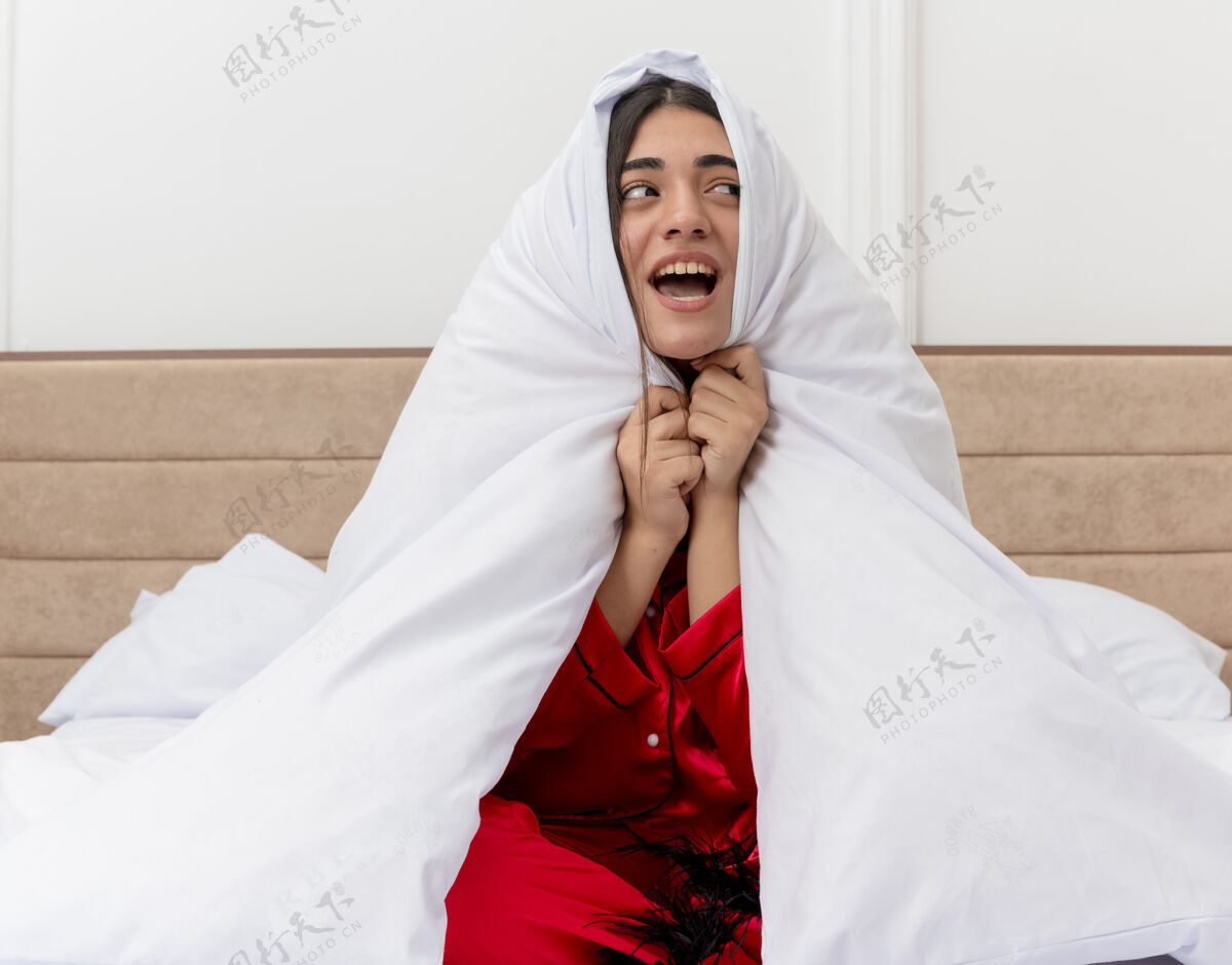 女人穿着红色睡衣的年轻漂亮女人坐在床上 裹着毛毯 开心地微笑着 看着卧室的内部室内包装年轻