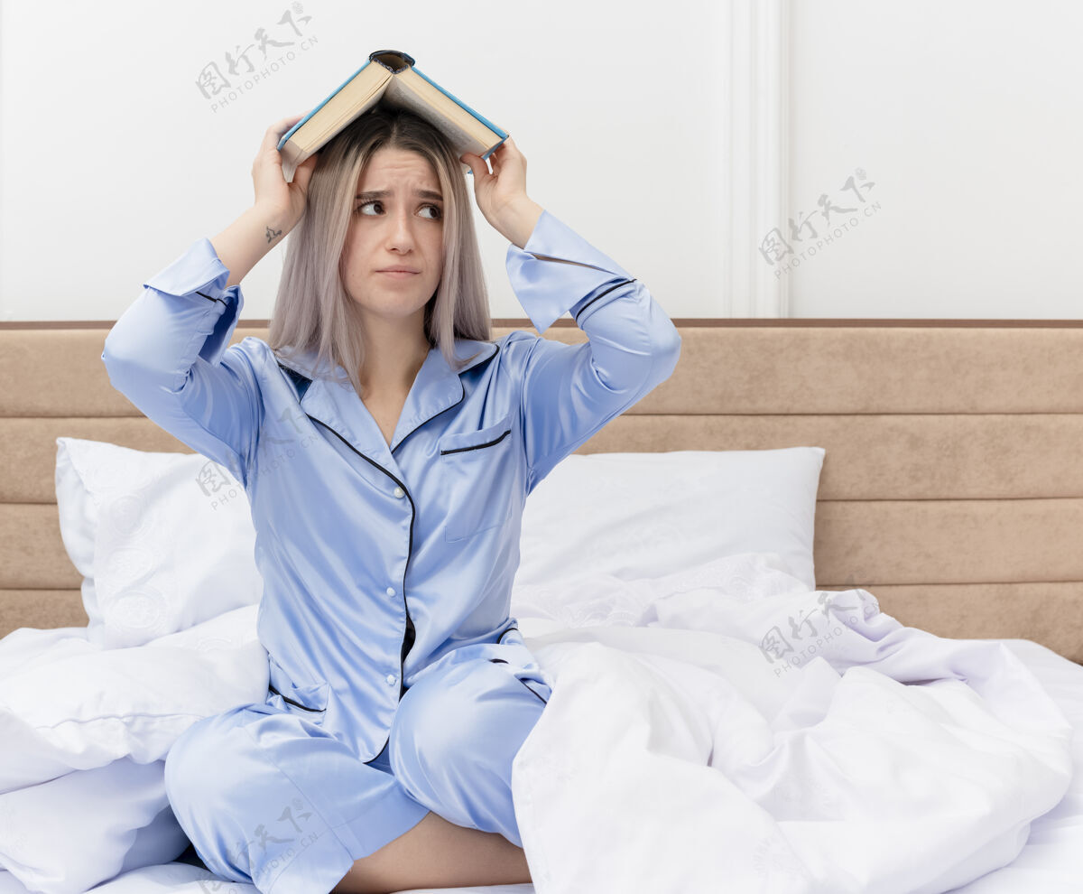 靠边穿着蓝色睡衣的年轻漂亮女人坐在床上 头上抱着书 在卧室里不满意地看着一边漂亮室内床