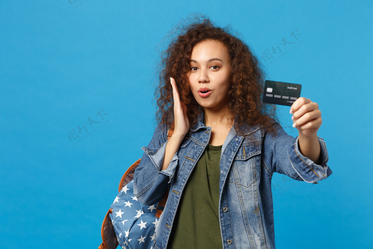 青少年年轻的非洲裔美国女孩十几岁的学生在牛仔服装 背包举行信用卡隔离在蓝色的墙上交换贷款钱