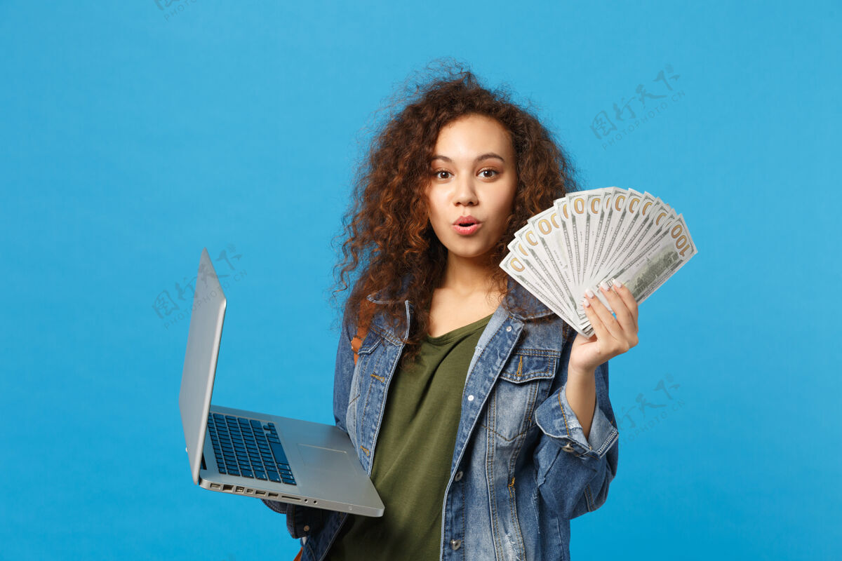 设备年轻的非洲裔美国女孩十几岁的学生在牛仔服装 背包举行个人电脑 球迷的现金钱隔离在蓝色的墙壁上使用人学生