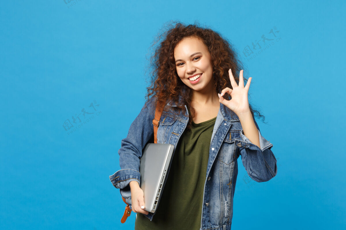 学生年轻的非洲裔美国女孩十几岁的学生在牛仔服装 背包举行电脑隔离在蓝色的墙壁上女性设备女孩