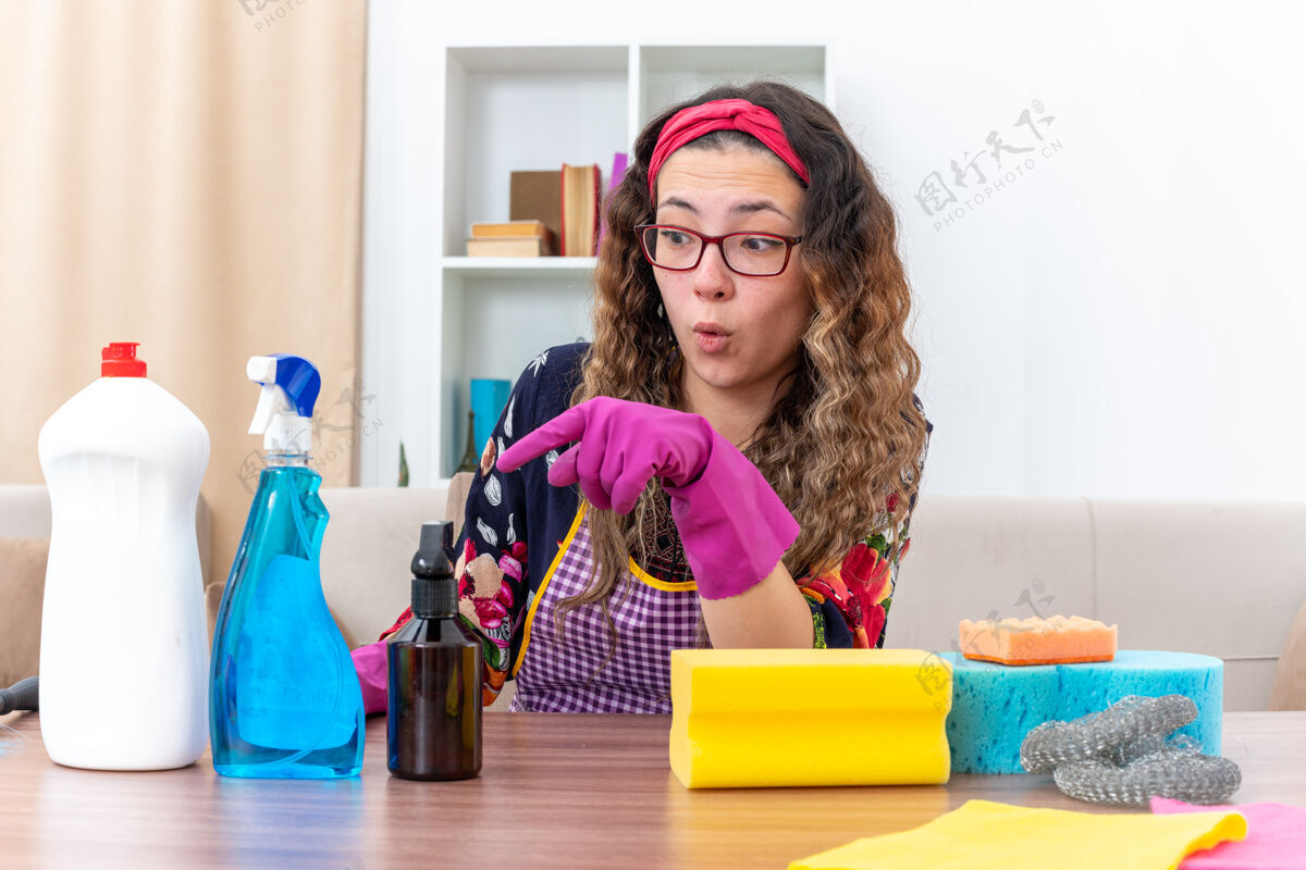 惊喜戴着橡胶手套的年轻女子坐在明亮的客厅里 手里拿着清洁用品和工具 看上去既惊讶又惊讶光清洁惊人