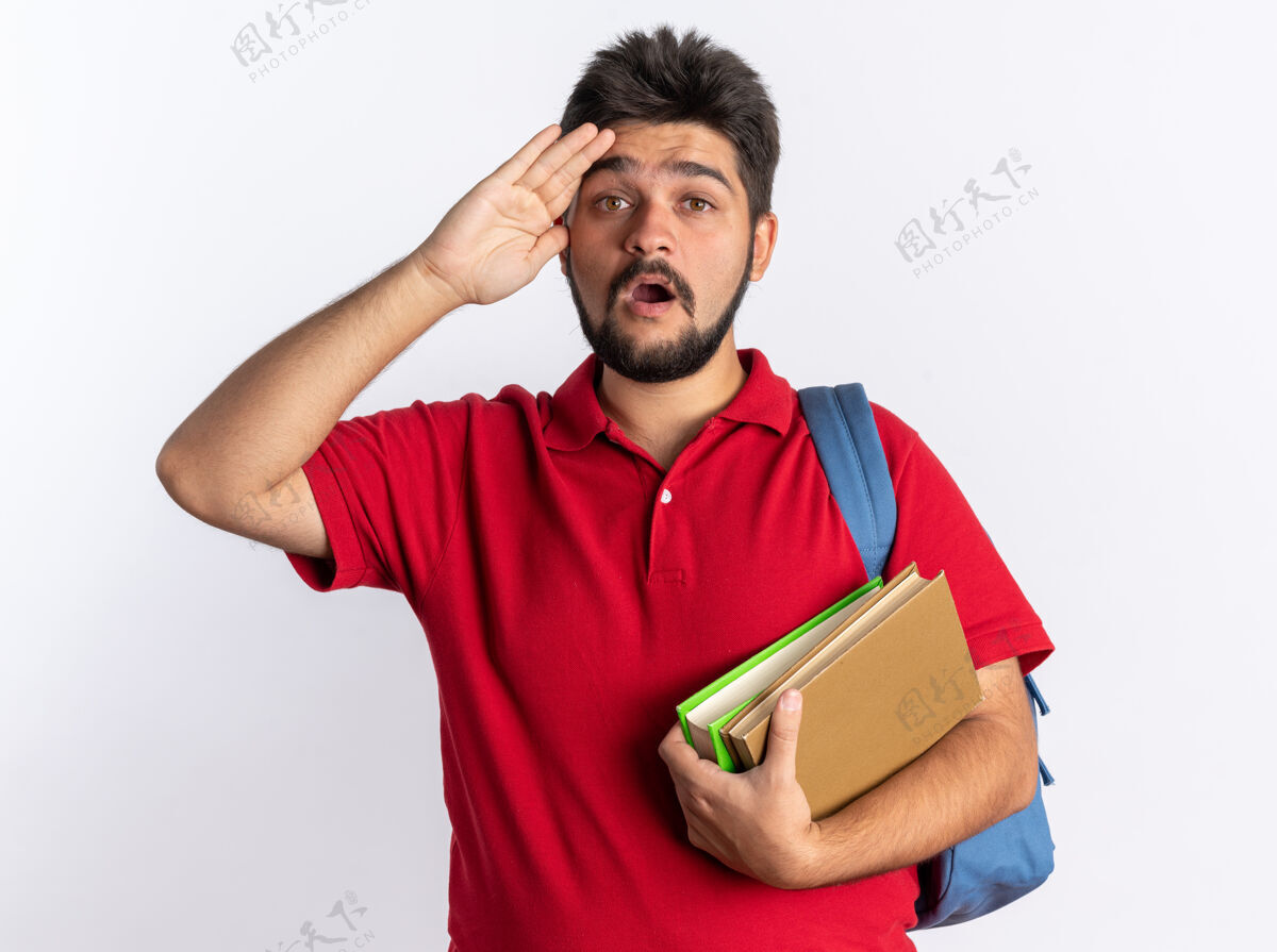 年轻年轻的留着胡子的学生 穿着红色马球衫 背着背包 手里拿着笔记本 站在白色的墙上 惊讶和惊讶胡须笔记本站