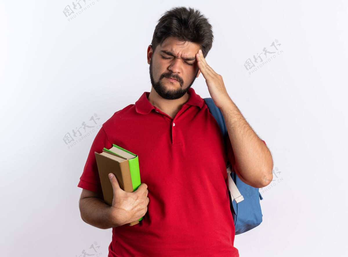 手势年轻的留着胡子的学生 穿着红色马球衫 背着背包 拿着笔记本 看起来不舒服 摸着太阳穴 站在白墙上感到疼痛触摸站立感觉