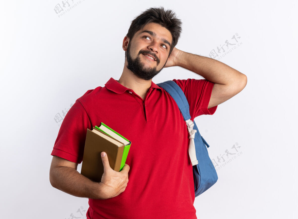 马球年轻的留着胡子的学生 穿着红色马球衫 背着背包 拿着笔记本 微笑着抬头 站在白色的墙上 思考着积极的事情积极手势思考