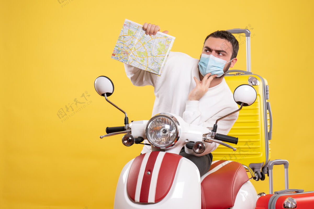 持有旅行概念的俯视图 困惑的家伙戴着医用面罩站在摩托车旁边 黄色手提箱放在上面 手里拿着医疗黄色高尔夫