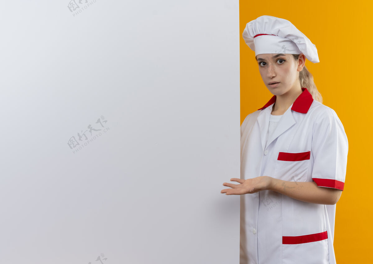 墙年轻漂亮的厨师穿着厨师制服站在白色的墙后 在橘色的墙上展示着被隔离的空手和复印空间印象年轻空