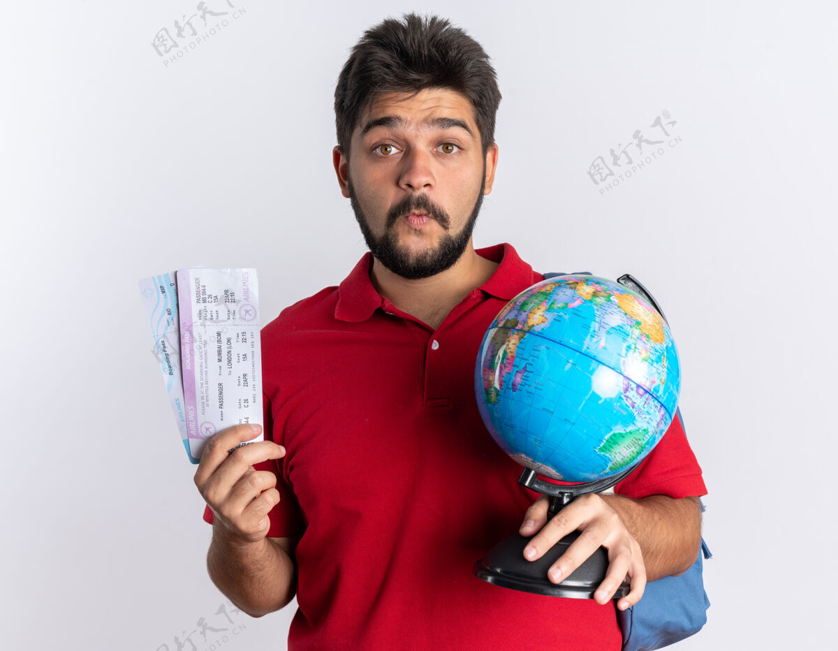 手势年轻的留着胡子的学生 穿着红色马球衫 背着背包 手里拿着机票 站在白色的墙上惊讶地看着地球仪地球仪惊喜背包