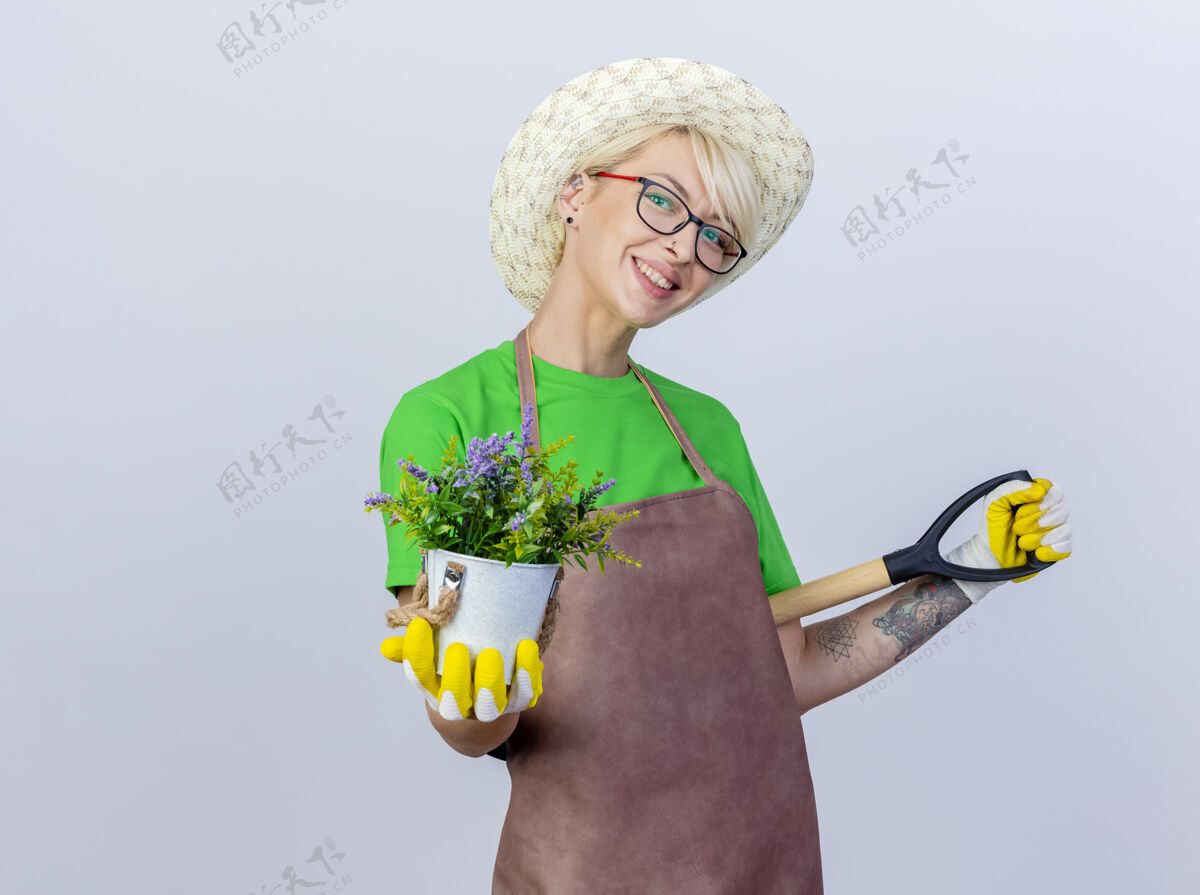 脸年轻的园丁 围裙短发 戴帽子 手里拿着铲子和盆栽 脸上带着微笑年轻抱帽子