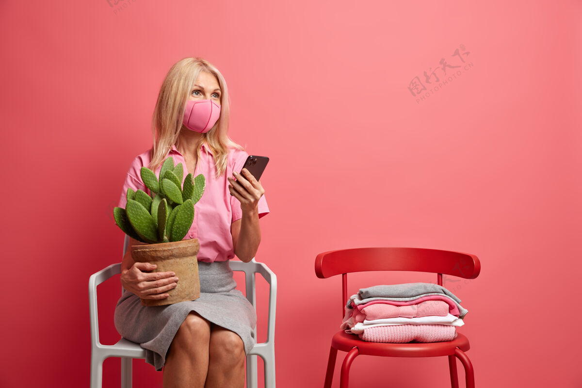 安全女人戴卫生口罩防止感染冠状病毒用智能手机聊天把一锅仙人掌摆在椅子上隔离在粉红色流行病检疫单独