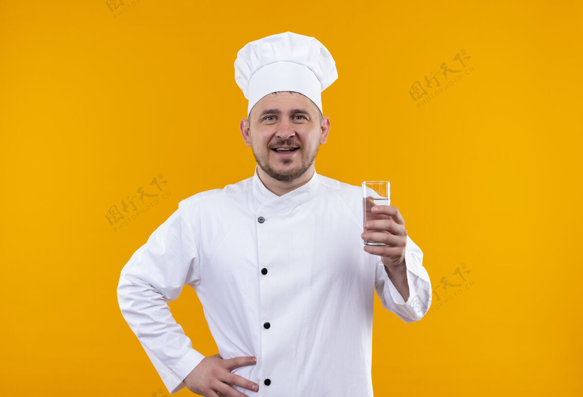烹饪快乐的年轻帅哥厨师身着厨师制服 手里拿着一杯孤立在橙色墙上的水手持厨师快乐