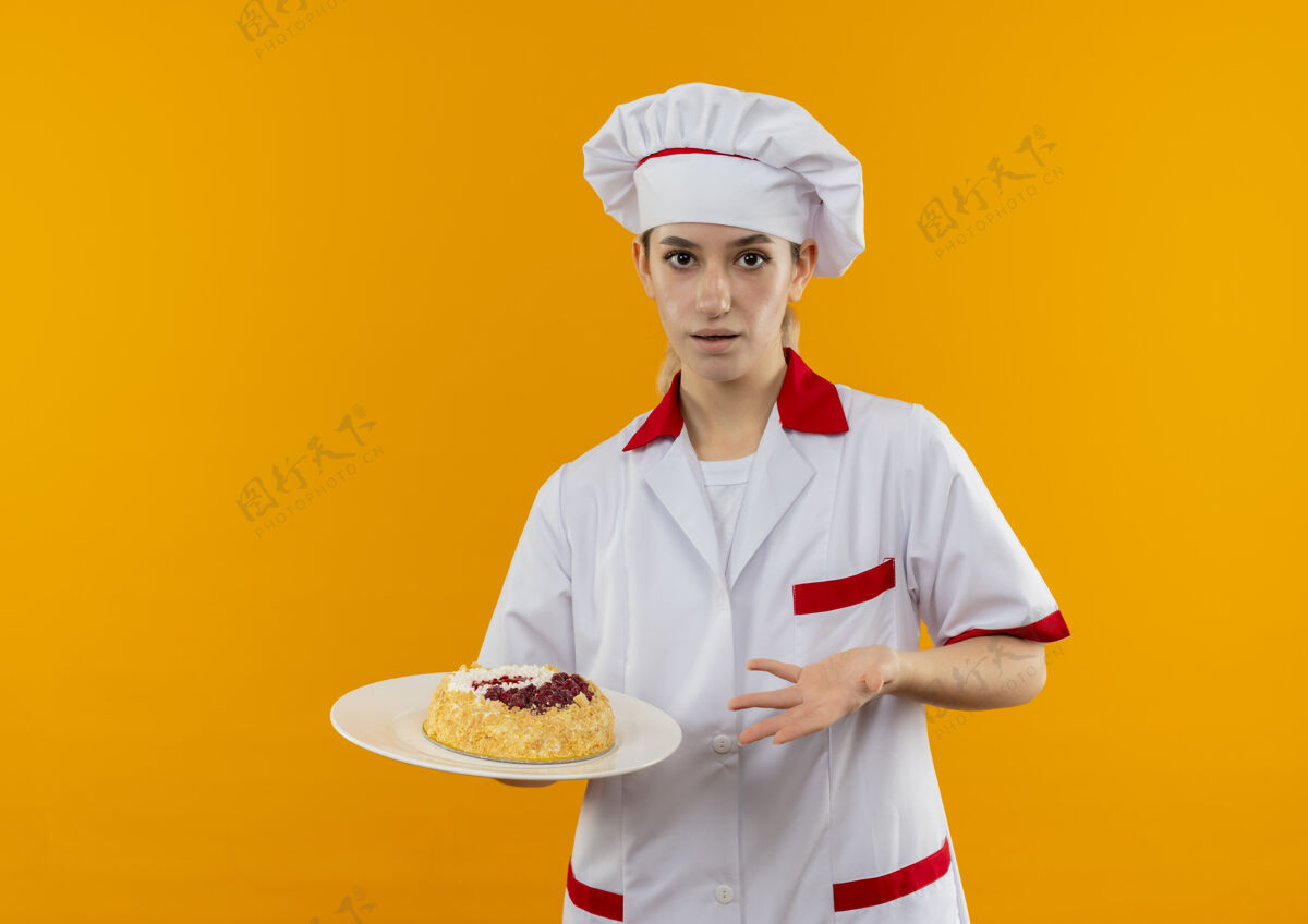 年轻穿着厨师制服的年轻漂亮厨师手拿并指着橘色墙上孤立的蛋糕盘厨师烹饪拿着