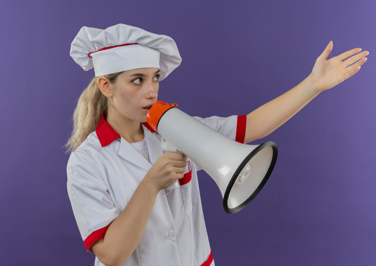 漂亮年轻漂亮的厨师穿着厨师制服 说话的人伸出手 看着紫色墙上孤立的一面壁板厨师伸展