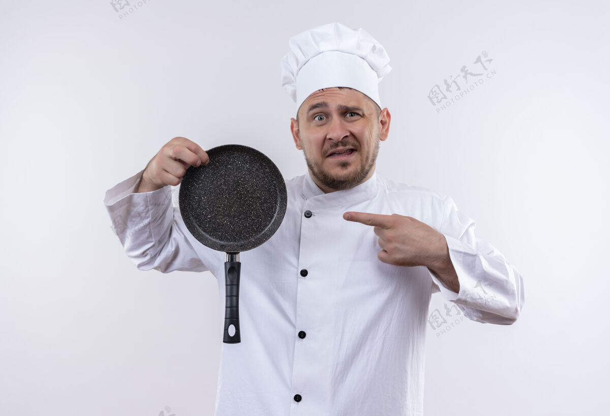 帅哥印象深刻的年轻帅哥厨师长制服举行 并指着煎锅隔离在白色的墙壁拿着指着薯条