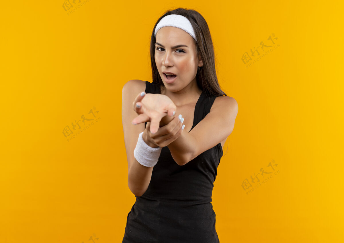 戴自信的年轻漂亮的运动女孩戴着头带和腕带伸出手和手臂隔离在橙色的墙上手臂运动自信