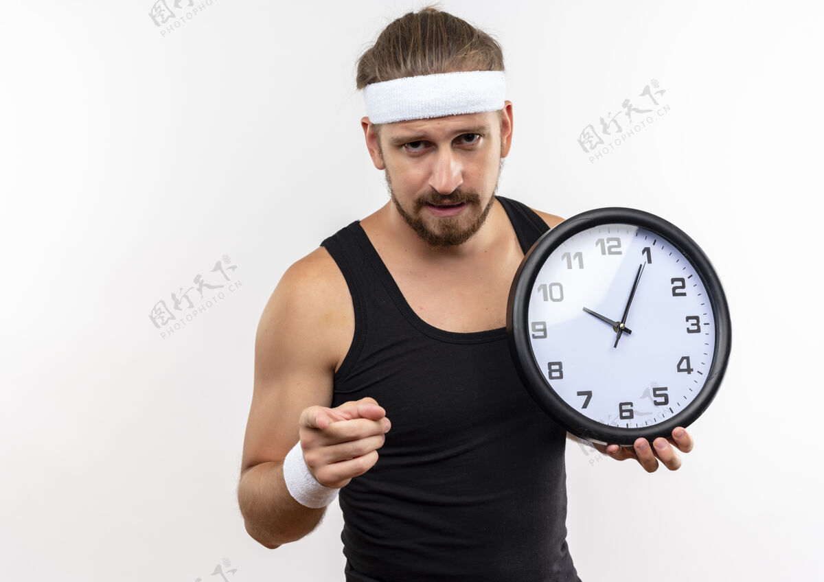 年轻自信的年轻帅气的运动型男人戴着头带和腕带拿着时钟 在白色的墙壁上孤立地指着复制空间英俊指着自信