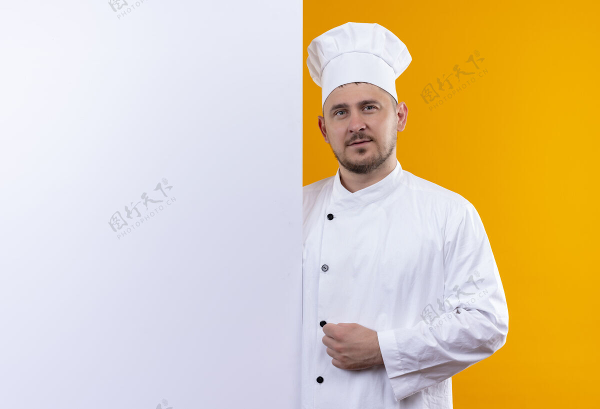 年轻自信的年轻帅哥厨师身着厨师制服站在白色的墙后 橙色的墙上有一个单独的复制空间墙壁厨师站立
