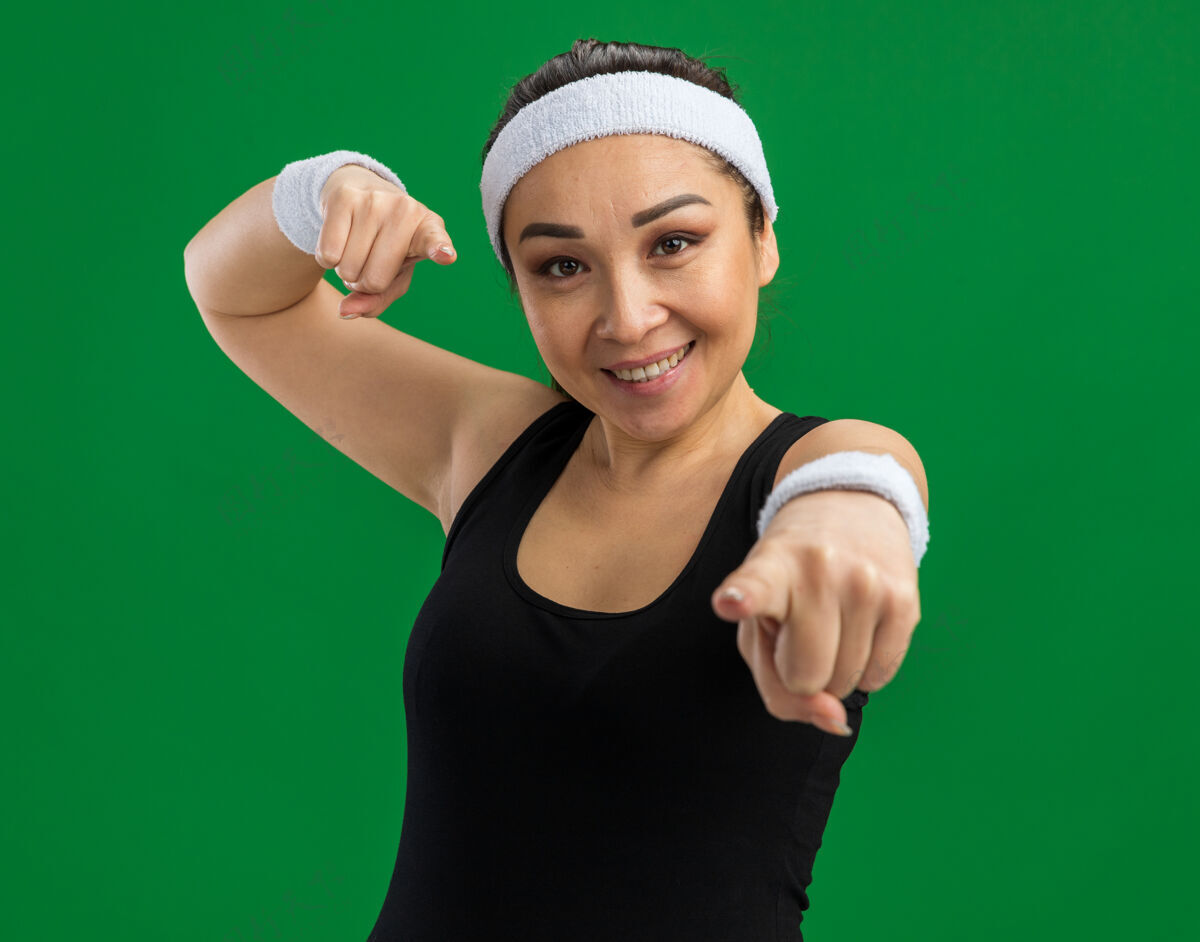 女人戴着头巾和臂章的年轻健身女士微笑着 用食指指着绿色的墙壁手指头带目录