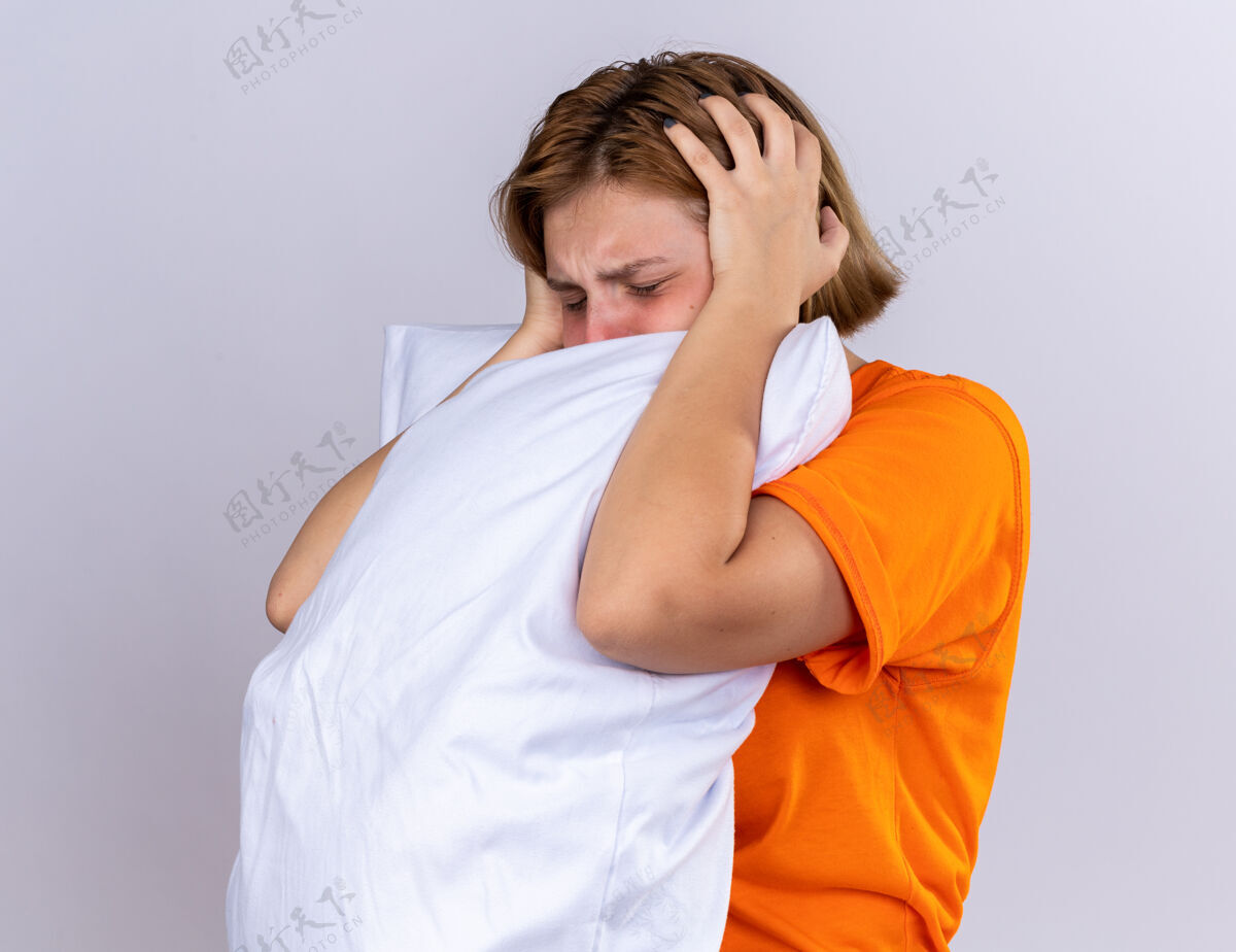 头痛不健康的年轻女子 穿着橙色t恤 抱着枕头 感觉不舒服 患流感 发烧 头痛 站在白墙上年轻感觉发烧