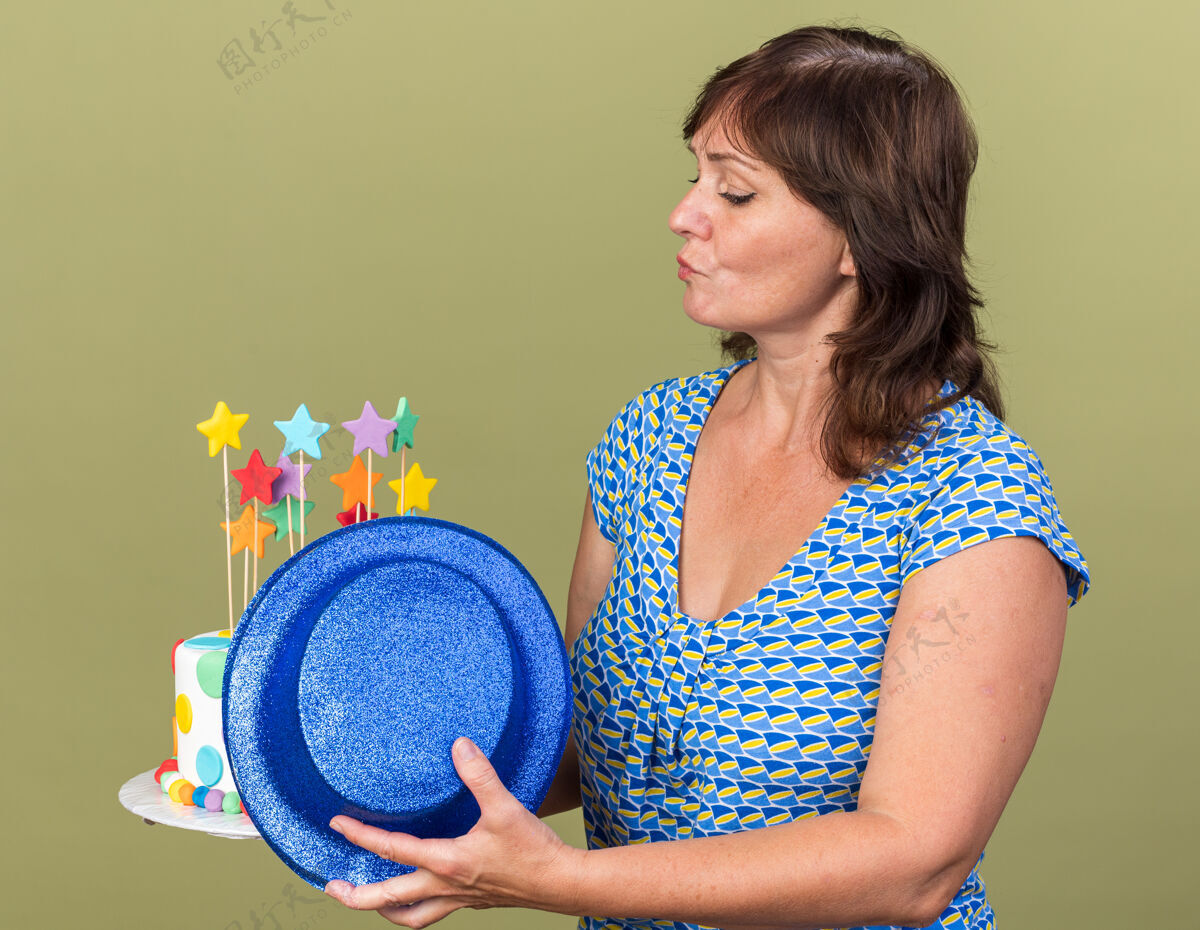 蛋糕中年妇女拿着生日蛋糕和派对帽 表情严肃自信地看着蛋糕 站在绿色的墙上庆祝生日派对表情中间信心
