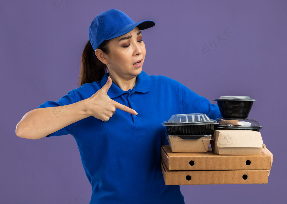 披萨身穿蓝色制服 头戴鸭舌帽的年轻送货员站在紫色的墙上 手里拿着比萨饼盒和食品包 用食指指着它们站着年轻人盒子