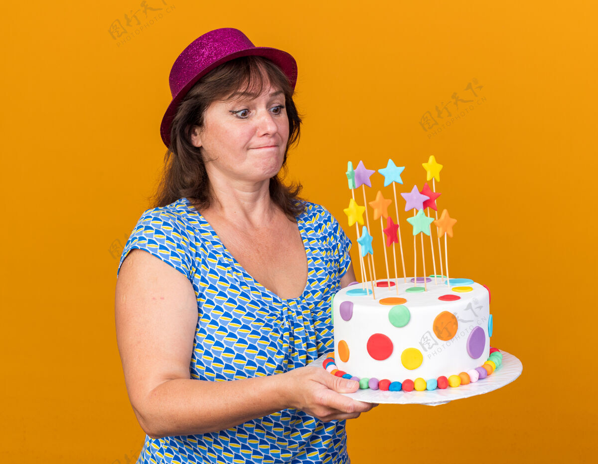 举行中年妇女戴着派对帽 拿着生日蛋糕 表情困惑地看着它 站在橘色的墙上庆祝生日派对年龄女人困惑