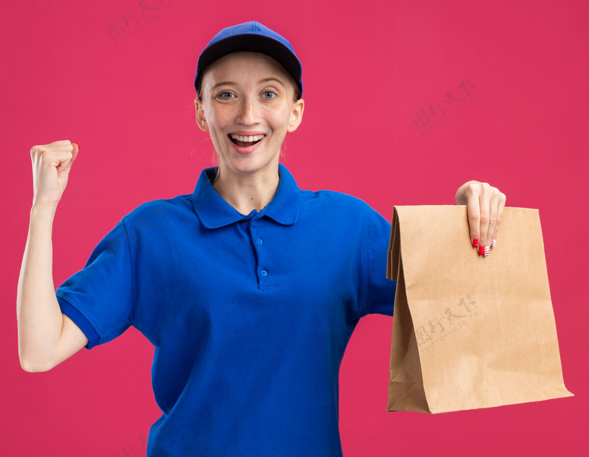 兴奋身穿蓝色制服 头戴鸭舌帽的年轻送货员手拿纸包 高兴而兴奋地握紧拳头站在粉红色的墙上举行交货女孩
