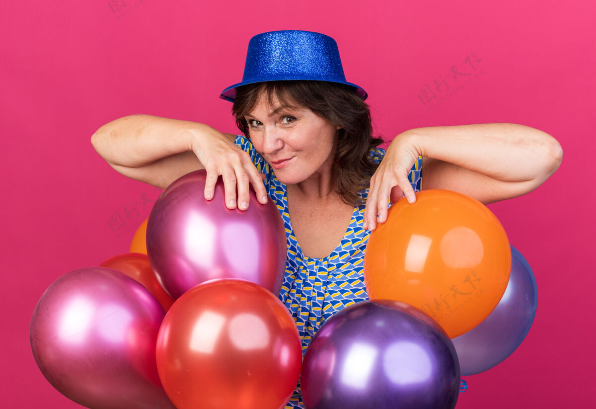 女人中年妇女戴着派对帽 手里拿着五颜六色的气球 幸福快乐地微笑着站在粉色的墙上庆祝生日派对中间生日微笑