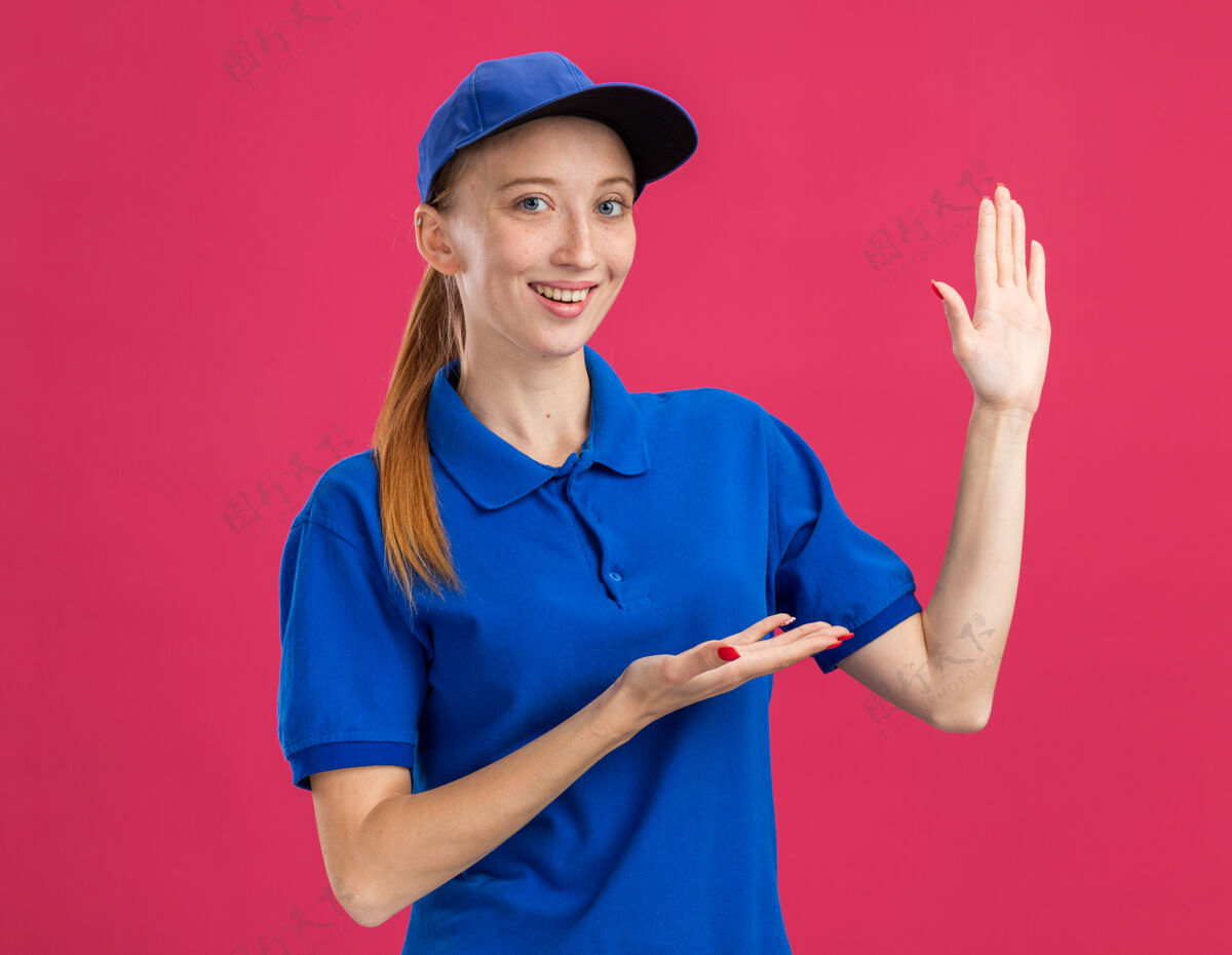 手臂身穿蓝色制服 头戴帽子的年轻送货女孩微笑着自信地站在粉色墙壁上展示复印空间制服交货年轻