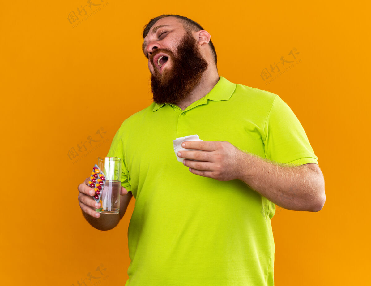 药丸一个留着不健康胡须 穿着黄色马球衫的男人 手里拿着一杯水和药片 感觉冷得要命 站在橙色的墙上打喷嚏感冒玻璃感觉