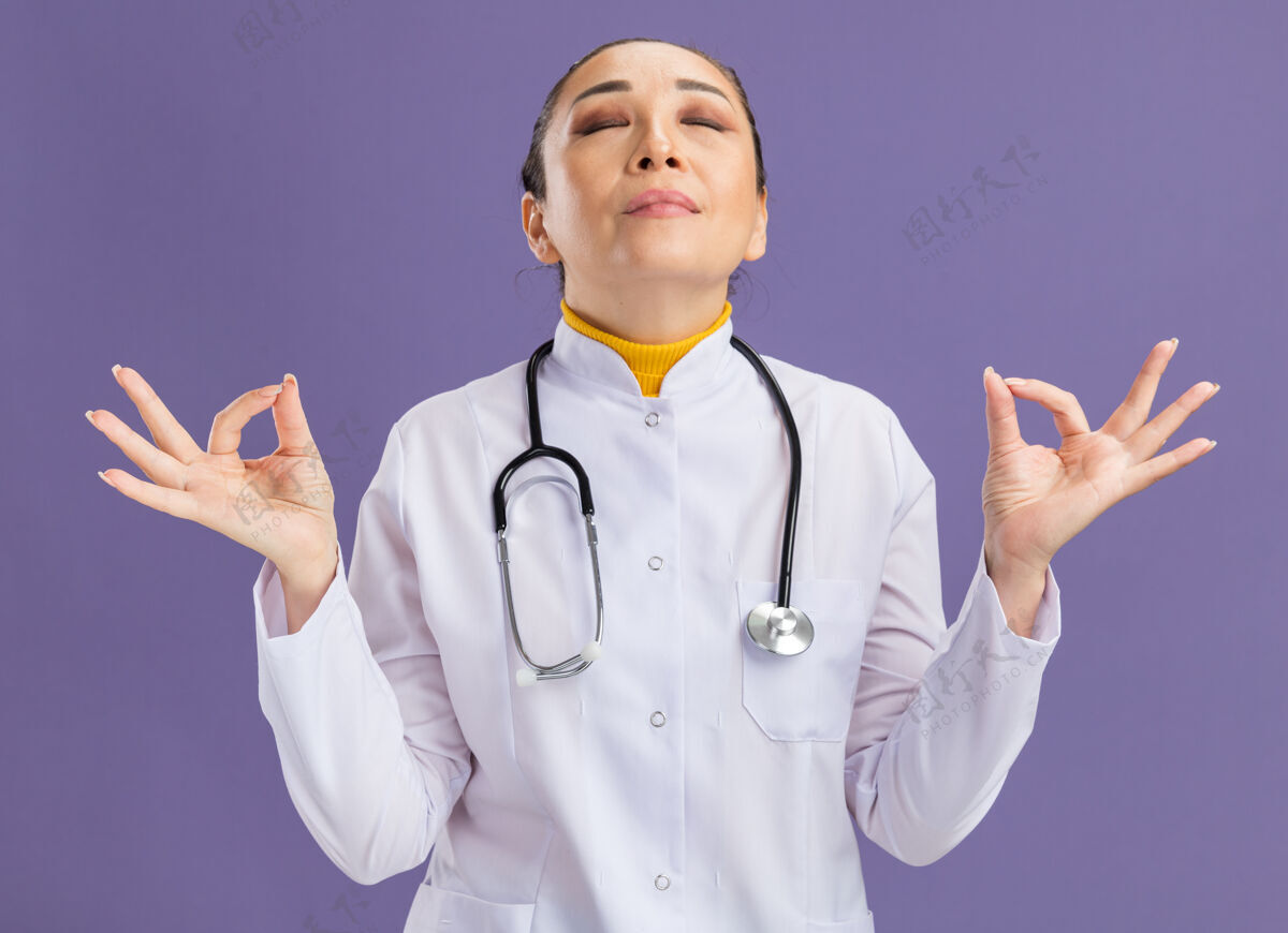 放松年轻的女医生穿着白色药衣 脖子上戴着听诊器 闭着眼睛做着冥想的手势 站在紫色的墙上放松手指做女人