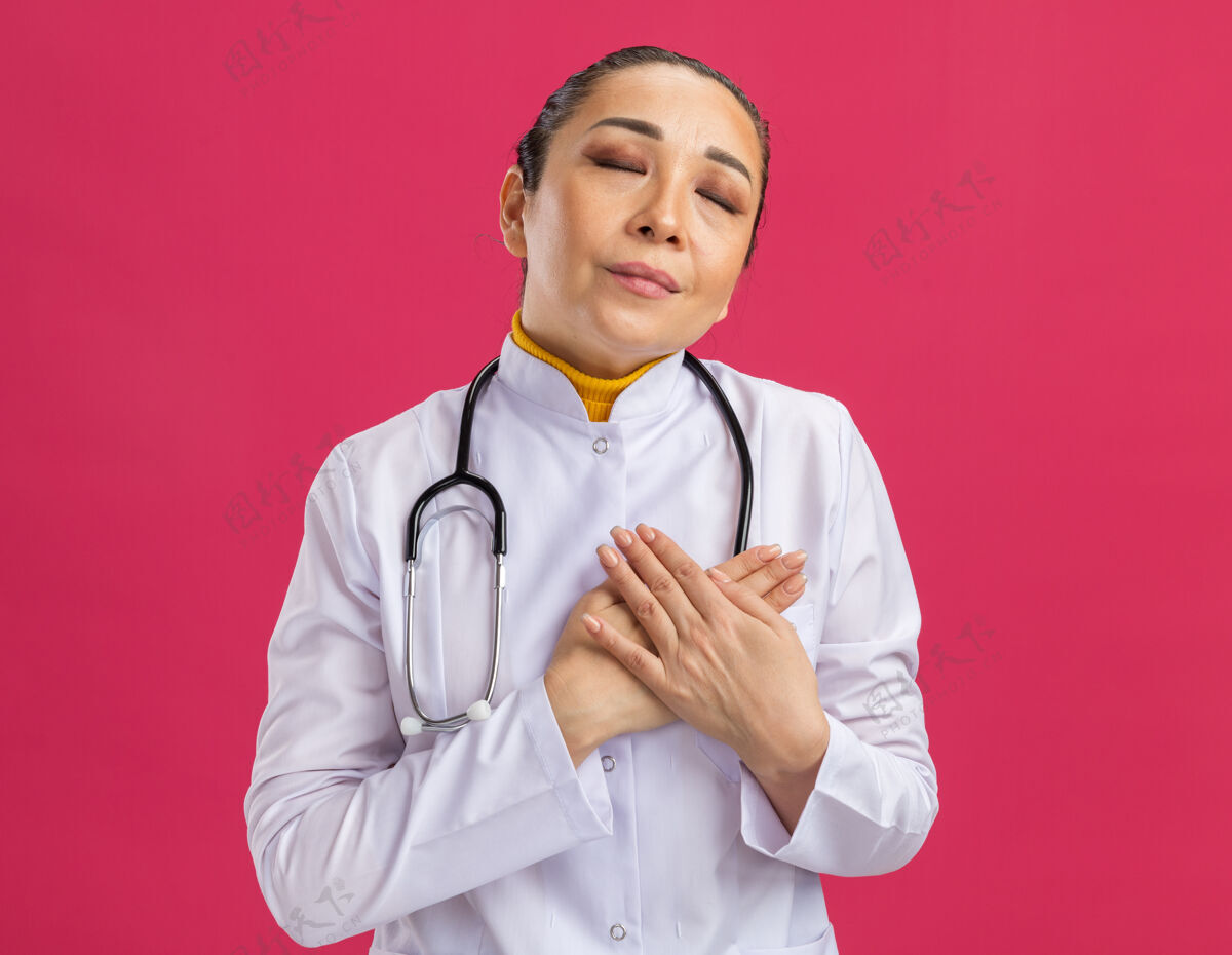 感情身穿白色药衣的年轻女医生 脖子上戴着听诊器 手放在胸前 站在粉红色的墙上 心怀感激年轻人抱着脖子