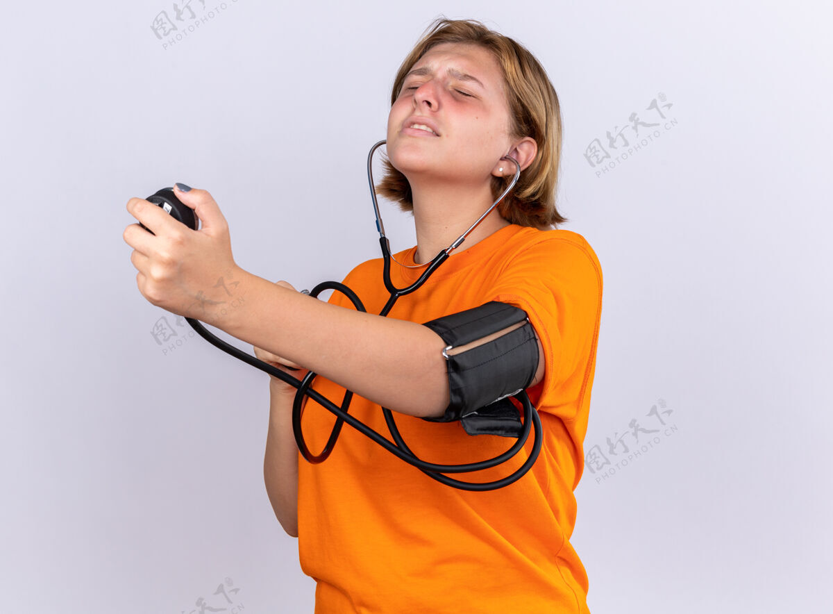 年轻不健康的年轻女子穿着橙色t恤 感觉不舒服 用血压计测量血压 站在白色的墙上 看起来很担心感觉血压计压力