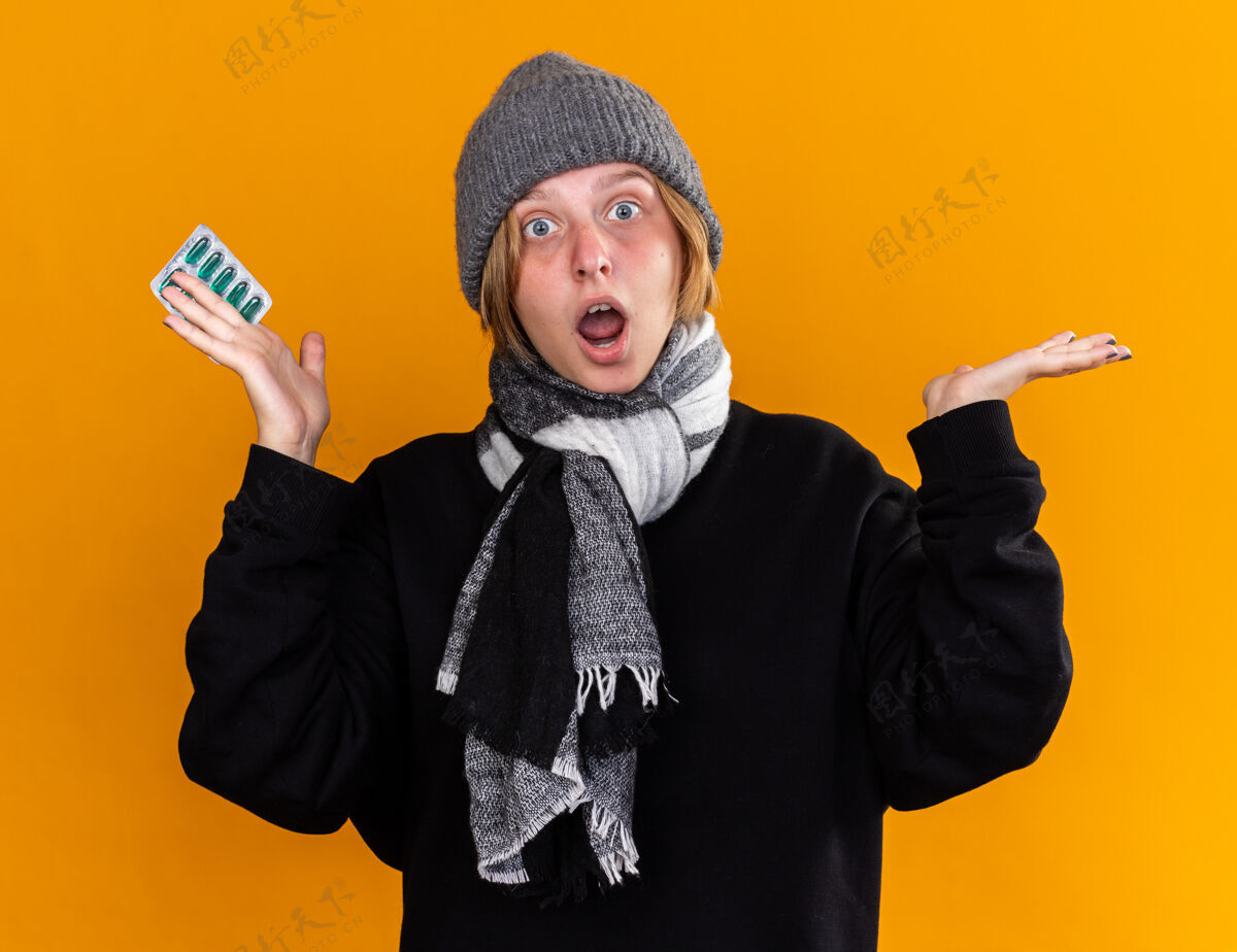 感冒不健康的年轻女子戴着暖和的帽子 脖子上围着围巾 感觉不舒服 患了感冒 吃了感冒药 站在橘色的墙上 看上去既惊讶又困惑惊人抱站