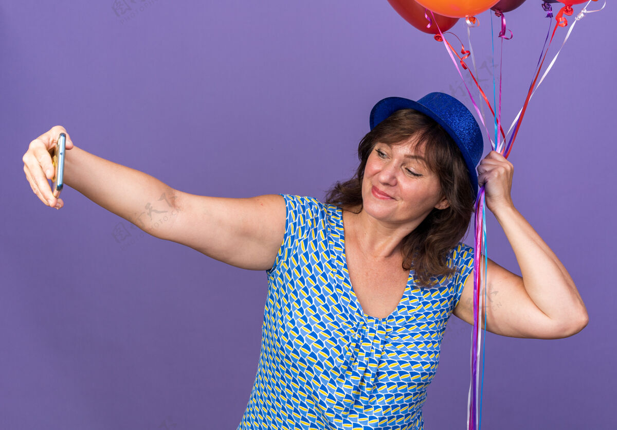 立场中年妇女戴着派对帽 手里拿着一束五颜六色的气球 开心地用智能手机自拍 站在紫色的墙上庆祝生日派对中间举行手机