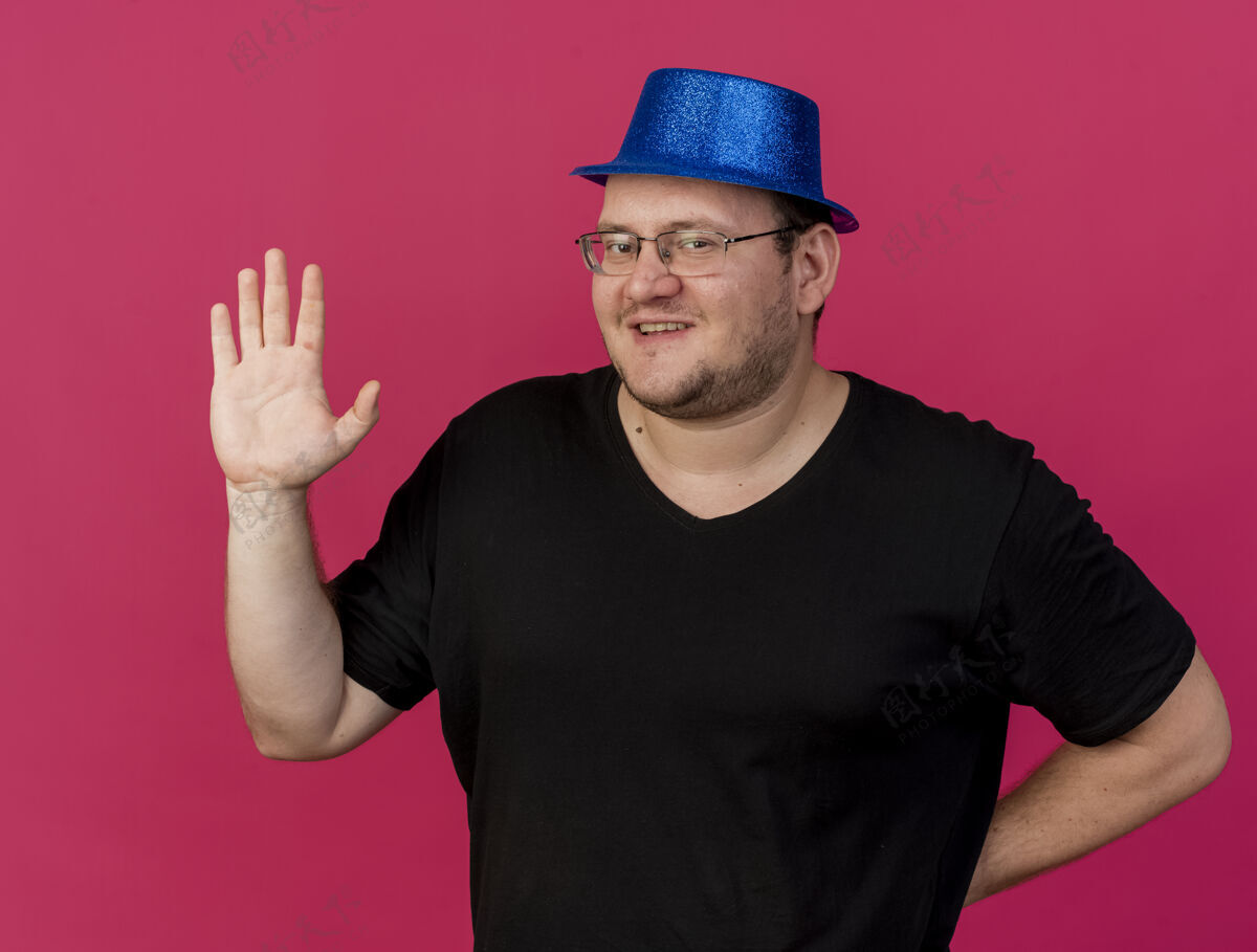 聚会微笑的成年斯拉夫人戴着眼镜 戴着蓝色派对帽 举起手站着举起男人站立