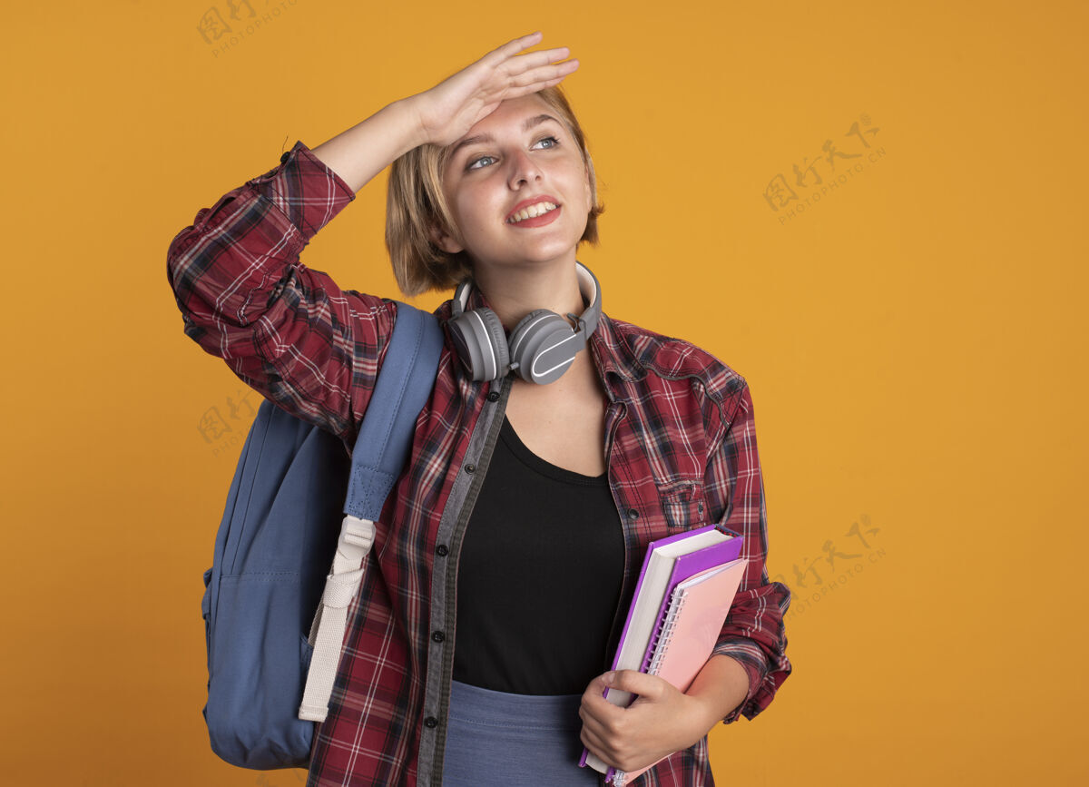 空间微笑的年轻斯拉夫女学生戴着耳机 背着背包 手掌放在额头 手里拿着书和笔记本额头学生耳机