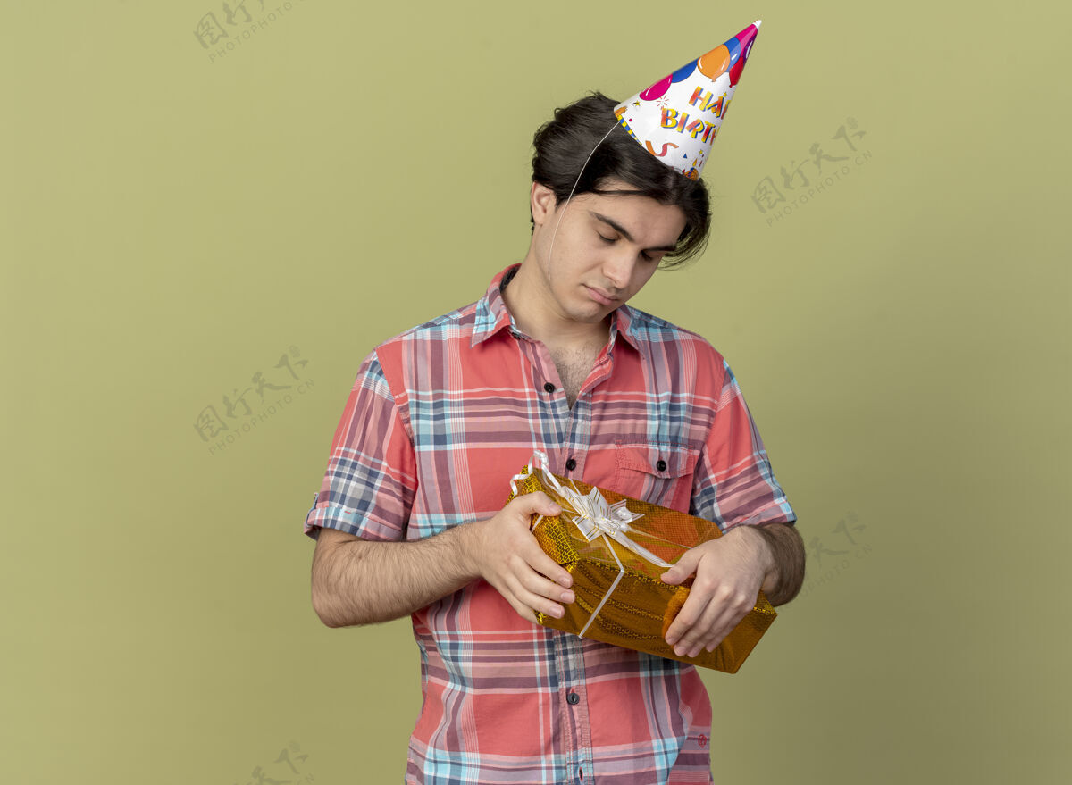 帅哥失望的白人帅哥戴着生日帽站在那里闭着眼睛拿着礼盒眼睛合上拿着