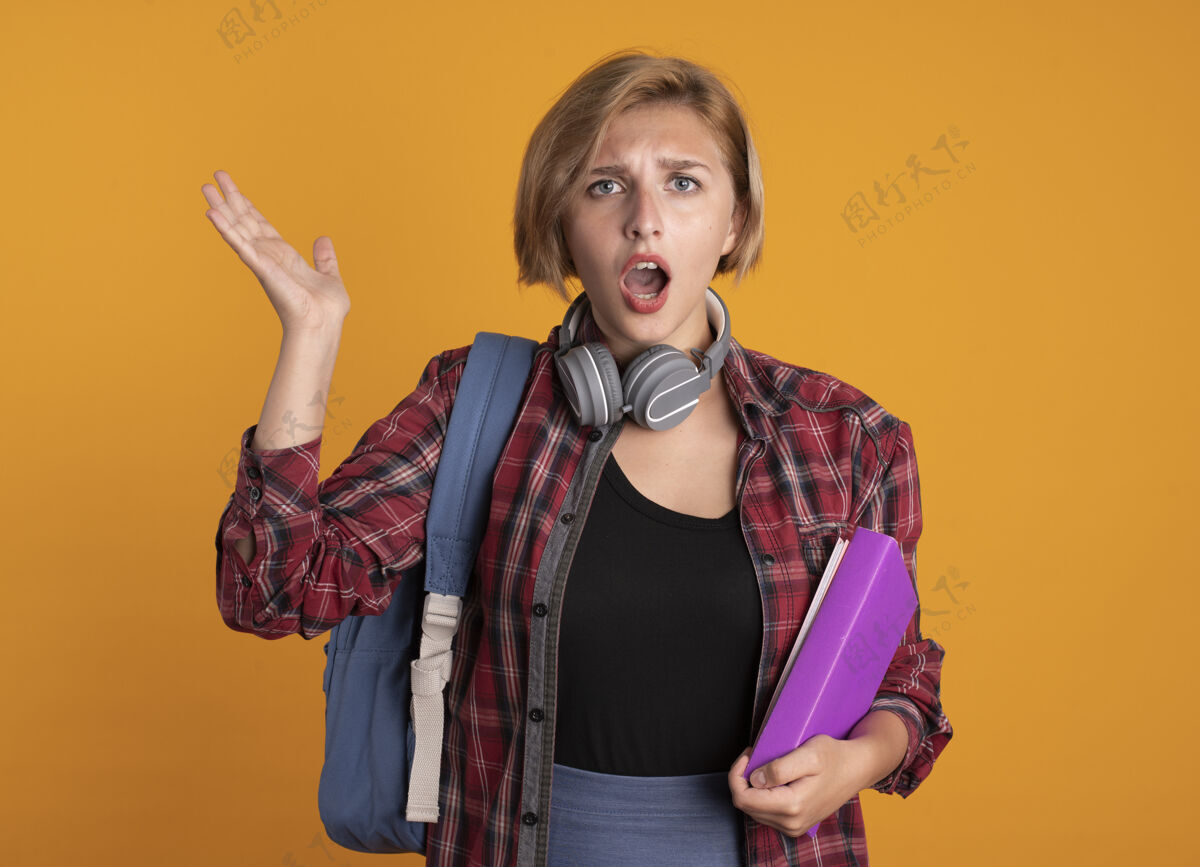 拿着无知的年轻斯拉夫学生女孩戴着耳机 背着书包 手举着书和笔记本学生笔记本橙色