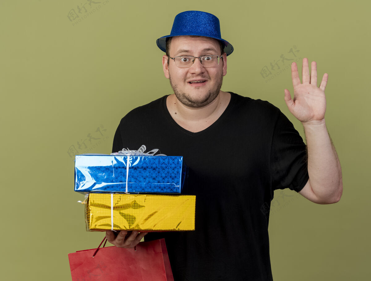 光学令人印象深刻的成年斯拉夫男子戴着眼镜 戴着蓝色的聚会帽 站着举起的手 拿着礼品盒和纸购物袋聚会提高站立