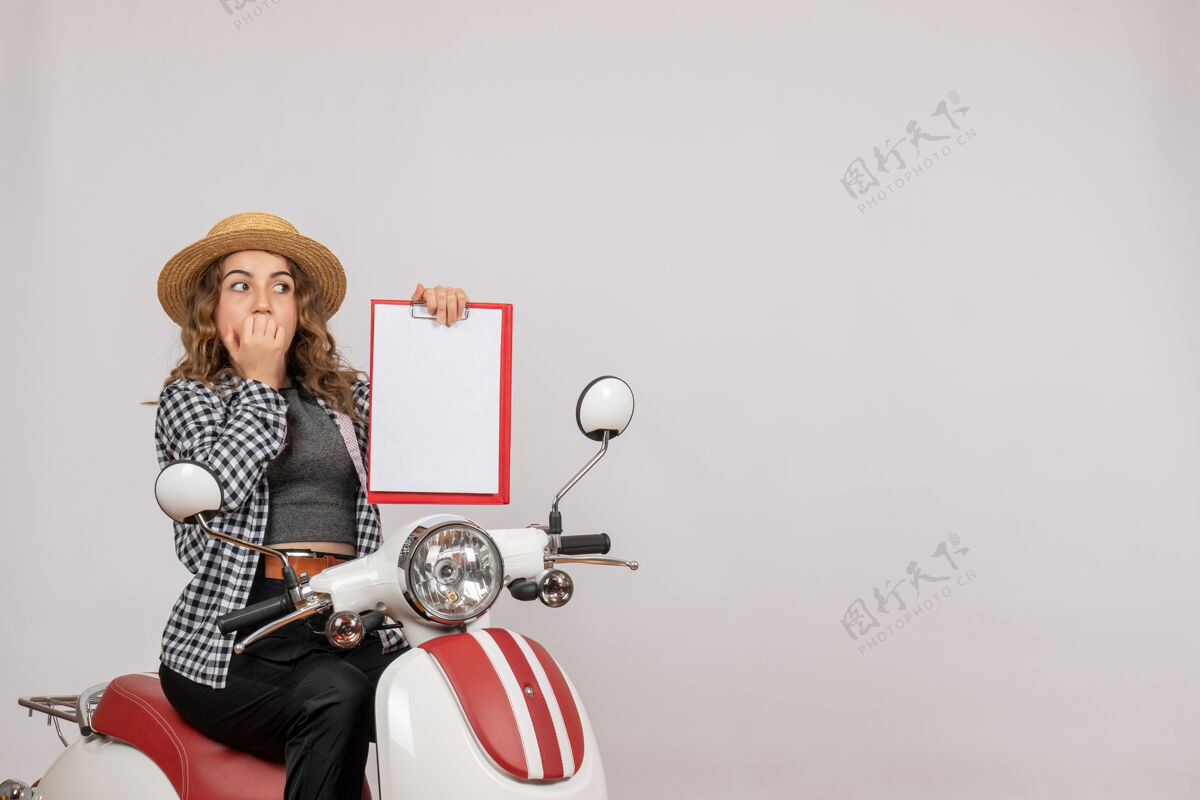 车辆坐在轻便摩托车上的迷惑不解的年轻女孩举着灰色的剪贴板人坐摩托车