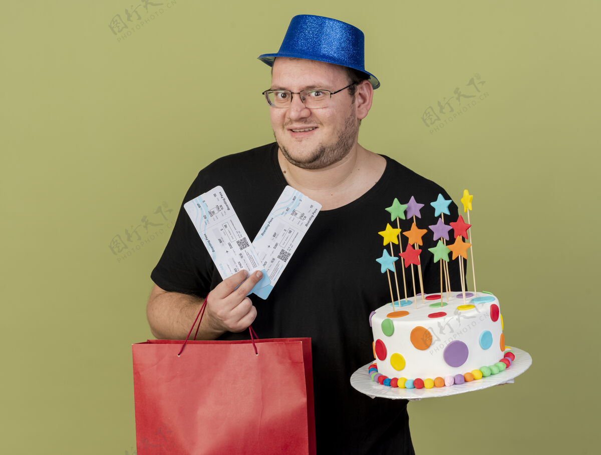 绿色兴奋的成年斯拉夫人戴着眼镜 戴着蓝色派对帽 手里拿着纸购物袋 生日蛋糕和机票举行包兴奋