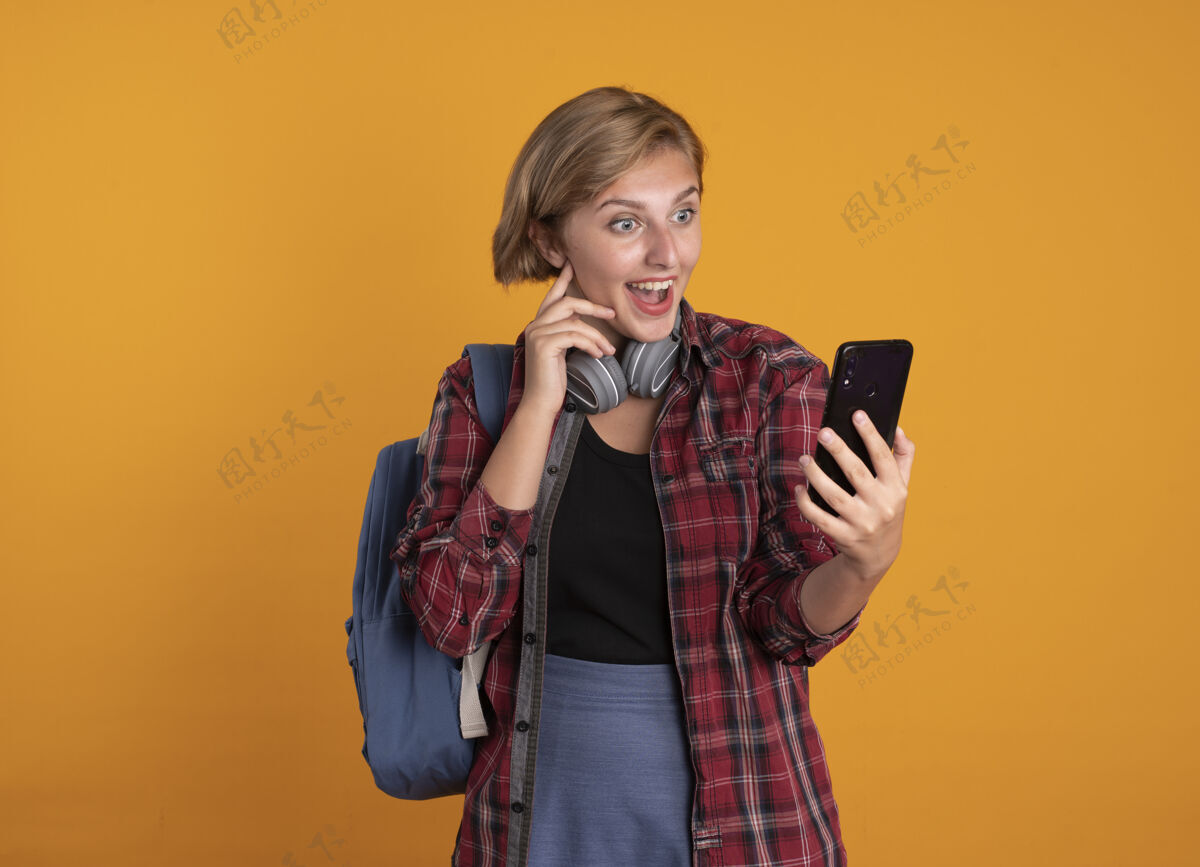 斯拉夫惊讶的年轻斯拉夫女学生戴着耳机背着背包把手放在脸上拿着手机看女孩复制橙色