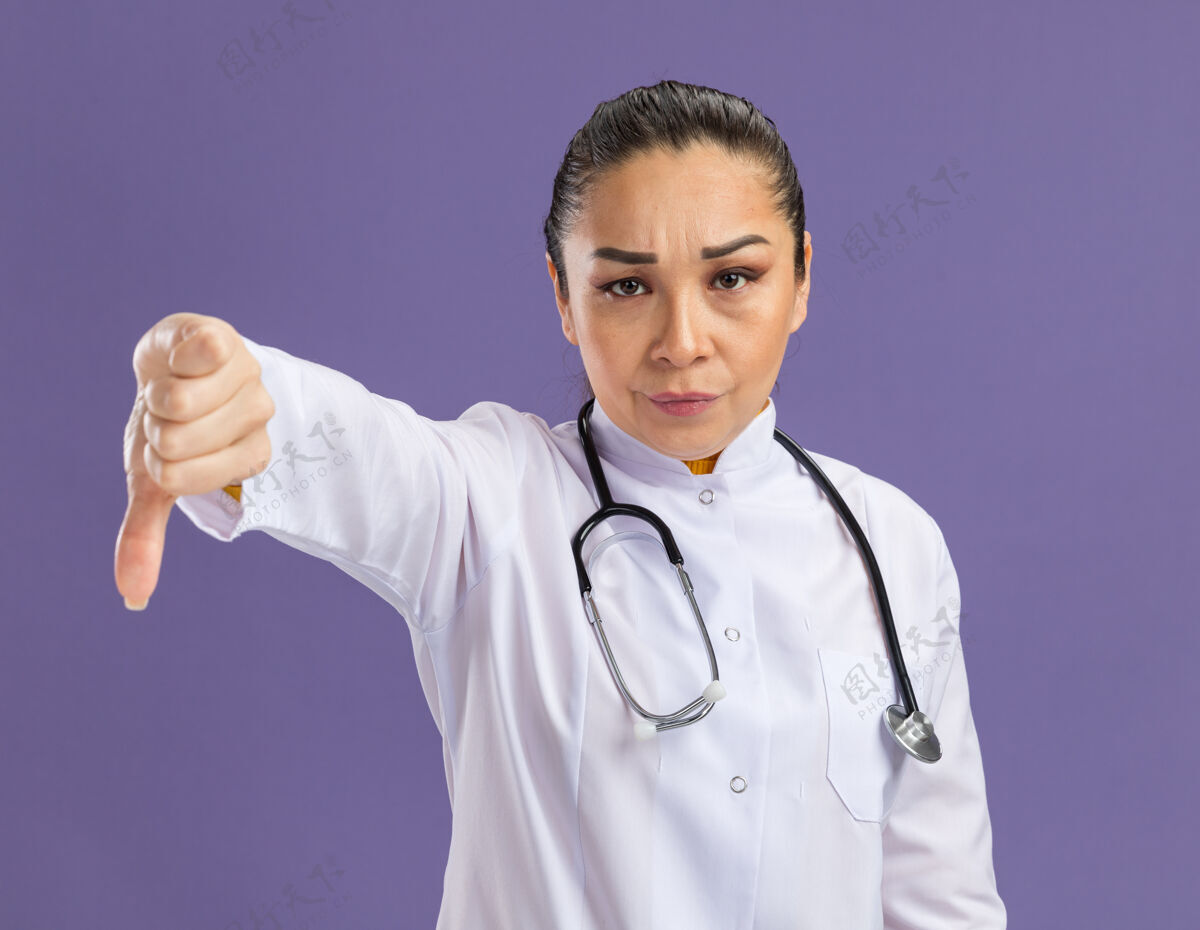 医生年轻的女医生 穿着白药衣 脖子上戴着听诊器 严肃的脸上竖着大拇指站在紫色的墙上站着围着听诊器