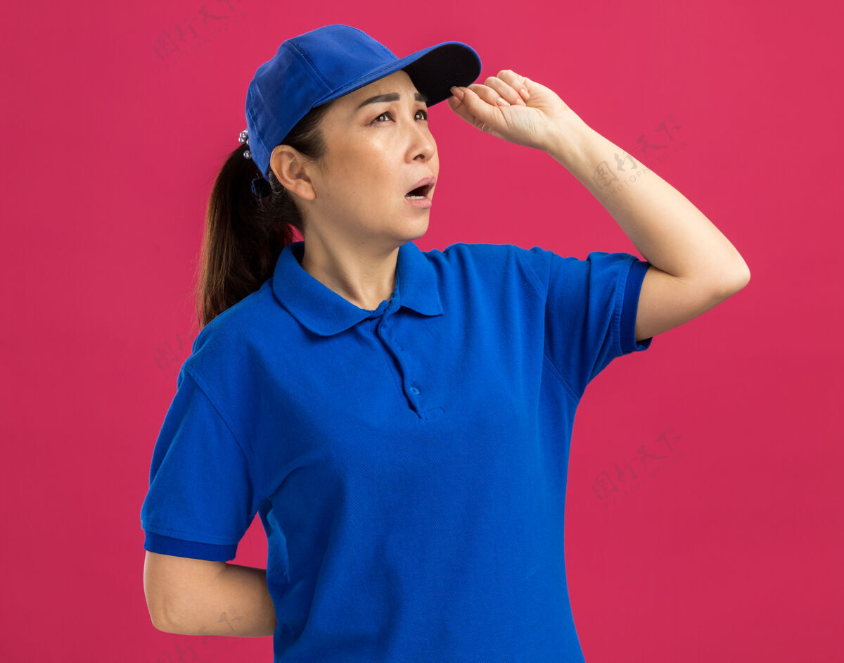 站立身穿蓝色制服 戴着帽子的年轻女送货员站在粉红色的墙上 一边沉思一边看着制服女人帽子